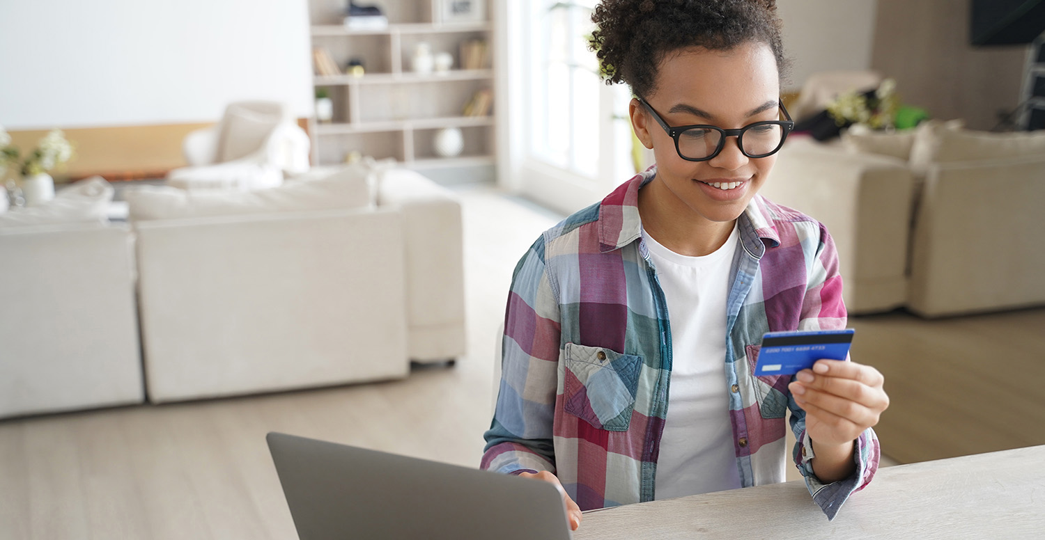 Teen girl holding banking card, shopping purchasing online, using laptop, making payment. Smiling teen paying using e-bank service, sitting at desk at home.